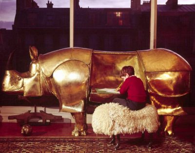 Large brass rhinoceros designed by Francois-Xavier Lalanne with folding sides being used as a desk by a little boy, also has a piggy-bank head with coin-drops in the ears --1967